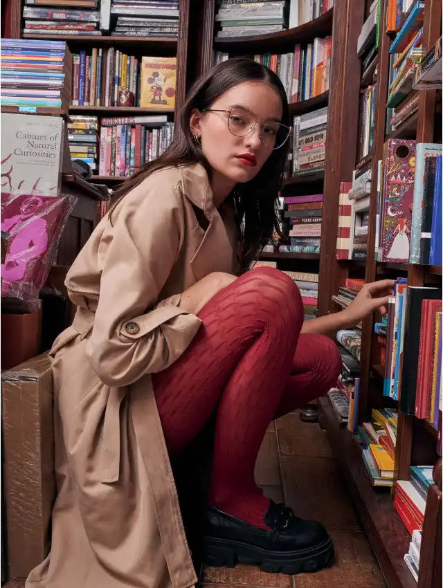 Donna con un trench beige, calze a fantasia rossa e loafers neri, accovacciata in una biblioteca.